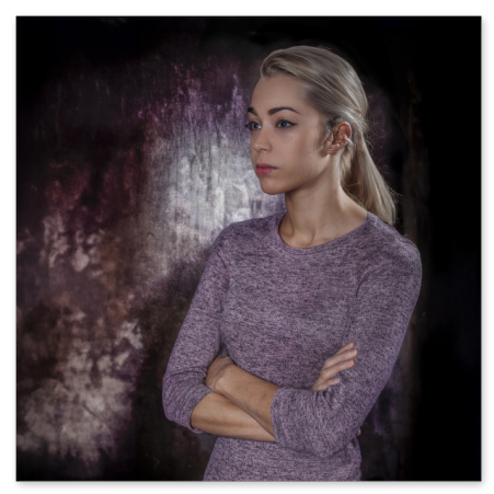 Eine junge Frau mit blondem, zurückgebundenem Haar steht mit verschränkten Armen in einem grauvioletten, eng anliegenden Strickkleid vor einem dunkel strukturierten Hintergrund und blickt nachdenklich zur Seite.
