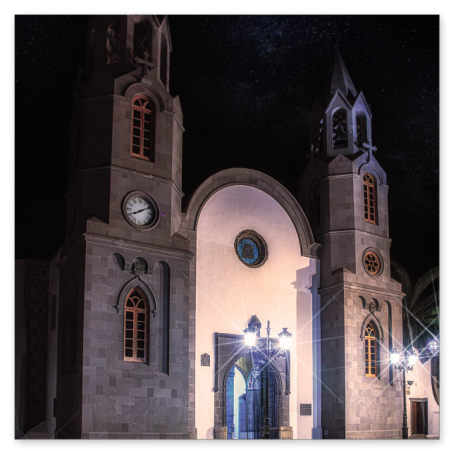  Eine hell erleuchtete, zweitürmige Kirche bei Nacht unter einem sternenklaren Himmel. Die neogotische Fassade aus dunklem Stein wird von Straßenlaternen und Bodenlichtern im Vordergrund angestrahlt.