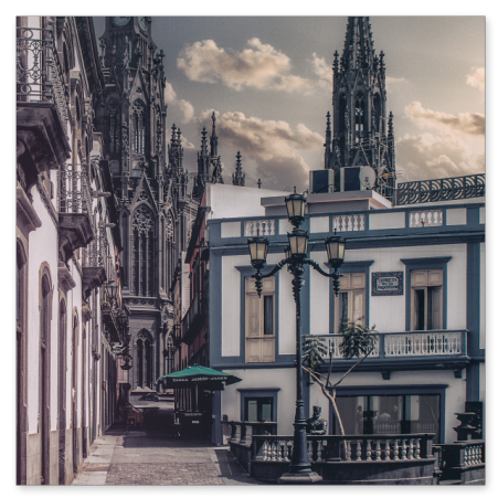 Blick durch eine historische Pflasterstraße in Arucas (Gran Canaria) auf die markanten, dunklen neogotischen Türme der Kirche San Juan Bautista unter leicht bewölktem Himmel.