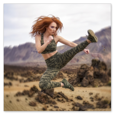 Eine sportliche, rothaarige Frau vollführt einen kraftvollen Sprungkick hoch in der Luft. Sie trägt ein olivgrünes Sport-Top und passende Leggings im Tarnmuster sowie Sneaker. Der Hintergrund zeigt eine karge Vulkan- oder Wüstenlandschaft mit Felsen.