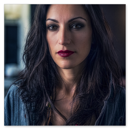 Nahaufnahme einer Frau mit langen, dunklen Haaren, braunen Augen und auffällig dunkelrotem Lippenstift. Sie trägt eine offene Jeansjacke, eine Kette aus Holzperlen und blickt direkt in die Kamera.
