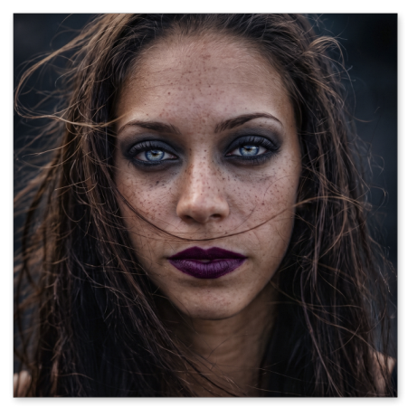 Enge Porträtaufnahme einer Frau mit zerzaustem, braunem Haar und ausgeprägten Sommersprossen. Sie fixiert die Kamera mit einem intensiven Blick aus hellen Augen, die von dunklem Lidschatten umrandet sind. Ihre Lippen sind in einem matten, dunklen Violett geschminkt.