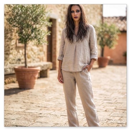 Eine junge Frau mit langen, braunen Haaren und dunklem Make-up steht auf dem sonnigen, gepflasterten Vorplatz eines mediterranen Steinhauses. Sie trägt eine locker sitzende beige Leinenbluse, eine passende Leinenhose sowie einfache Riemchensandalen.