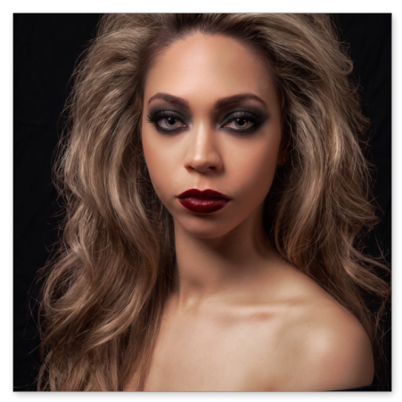 Frontales Porträt einer jungen Frau mit voluminösem, aschblondem Haar und intensivem Smokey-Eyes-Make-up. Sie trägt dunkelroten Lippenstift sowie ein schulterfreies schwarzes Oberteil vor schwarzem Hintergrund.