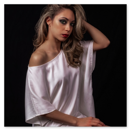 Eine junge Frau mit voluminösem, blondiertem Haar sitzt in einem fließenden, weißen Seidenkleid vor tiefschwarzem Hintergrund. Sie berührt mit einer Hand ihr Haar und blickt mit ausdrucksstarkem Make-up und roten Lippen zur Seite.