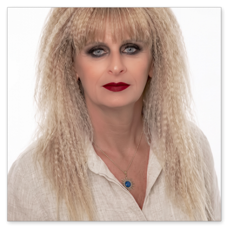 Frontales Porträt einer Frau mit blonden, gekreppten Haaren und Pony vor weißem Hintergrund. Sie trägt eine beige Leinenbluse, eine Halskette mit blauem runden Anhänger, dunkles Augen-Make-up und tiefroten Lippenstift.